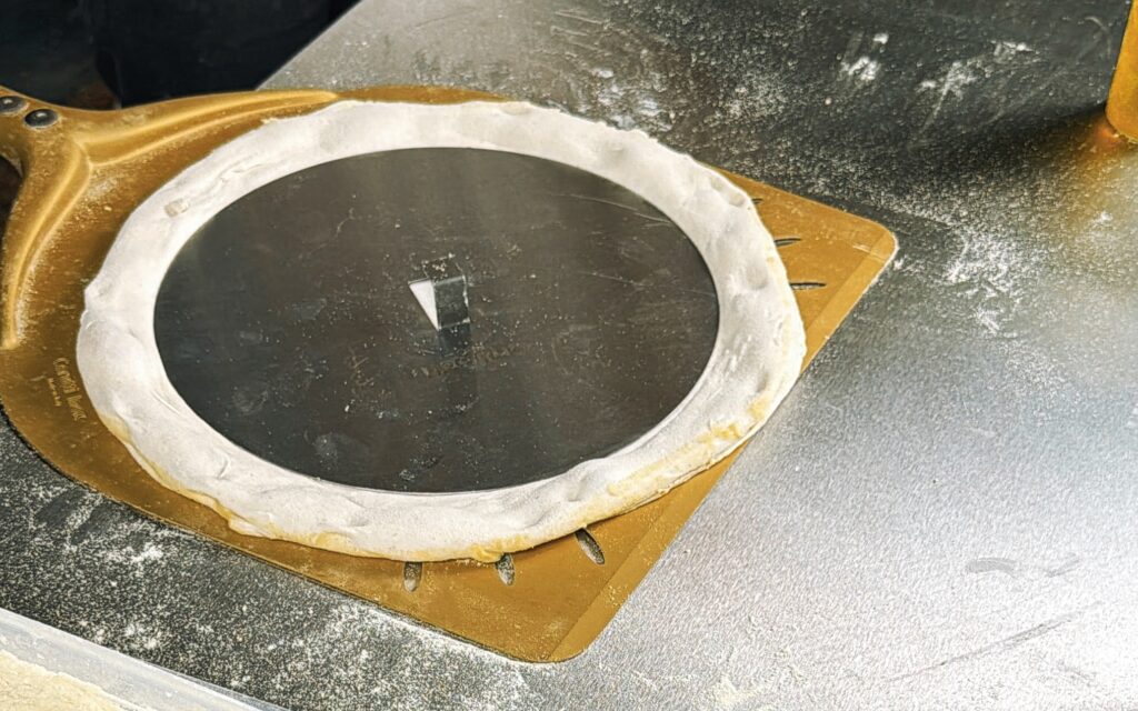 Teig auf der Pizzaschaufel, rundes Edelstahlblech liegt drauf – bereit für den Ofen