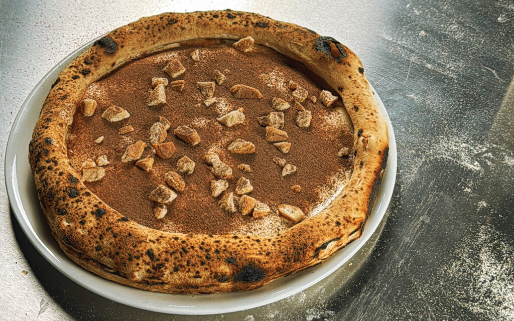 Fertige Tiramisu Pizza von oben – gleichmäßiger Kakaospiegel, grob gebrochene Biskotten, schöner Cornicione