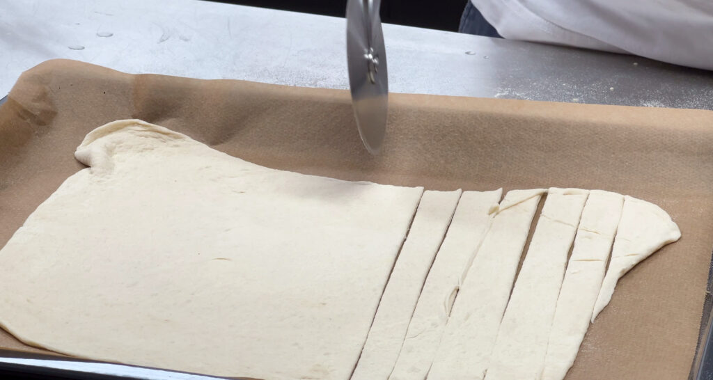 Dünn ausgerollter Pizzateig wird mit einem Pizzaschneider in Streifen geschnitten