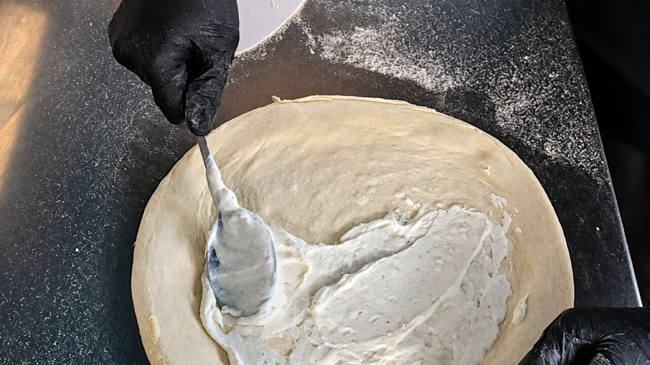 Weiße Ricotta-Sauce wird mit dem Löffel als Base auf den ausgezogenen Pizzateig gestrichen