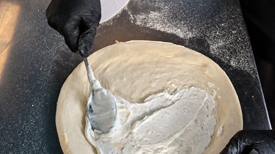 Weiße Ricotta-Sauce wird mit dem Löffel als Base auf den ausgezogenen Pizzateig gestrichen